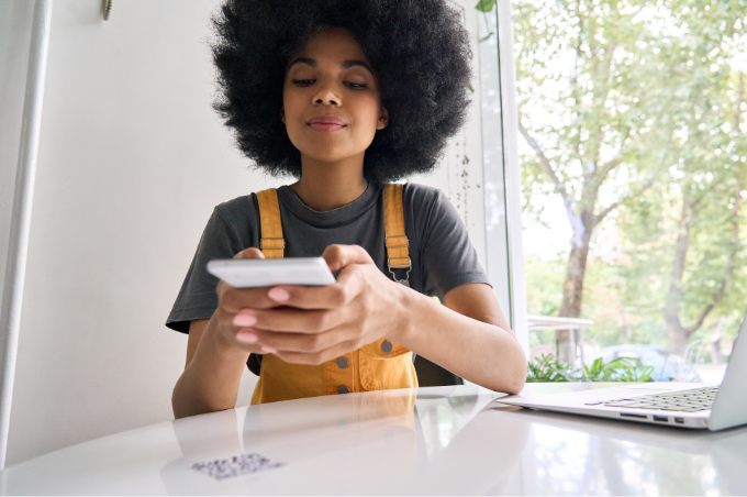 Eine junge Frau sitzt an einem Tisch
und bedient ihr Smartphone. Ihr
geöffneter Laptop steht auf dem
Tisch.