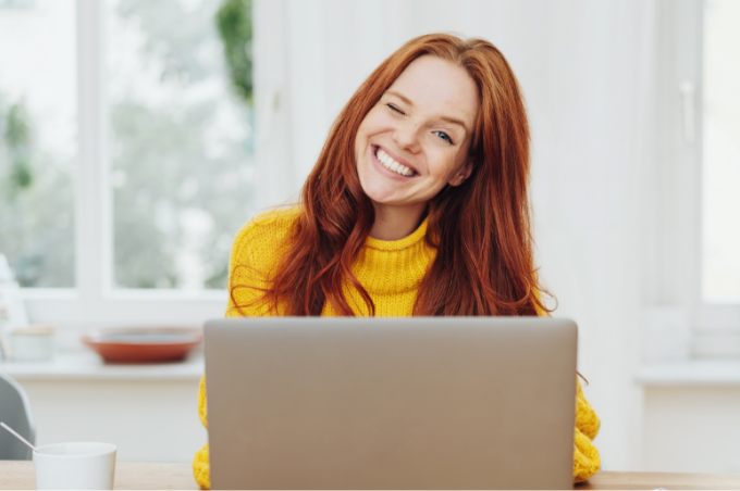 Eine junge Frau mit heiterem Lächeln
sitzt am Tisch vor ihrem geöffneten
Laptop.